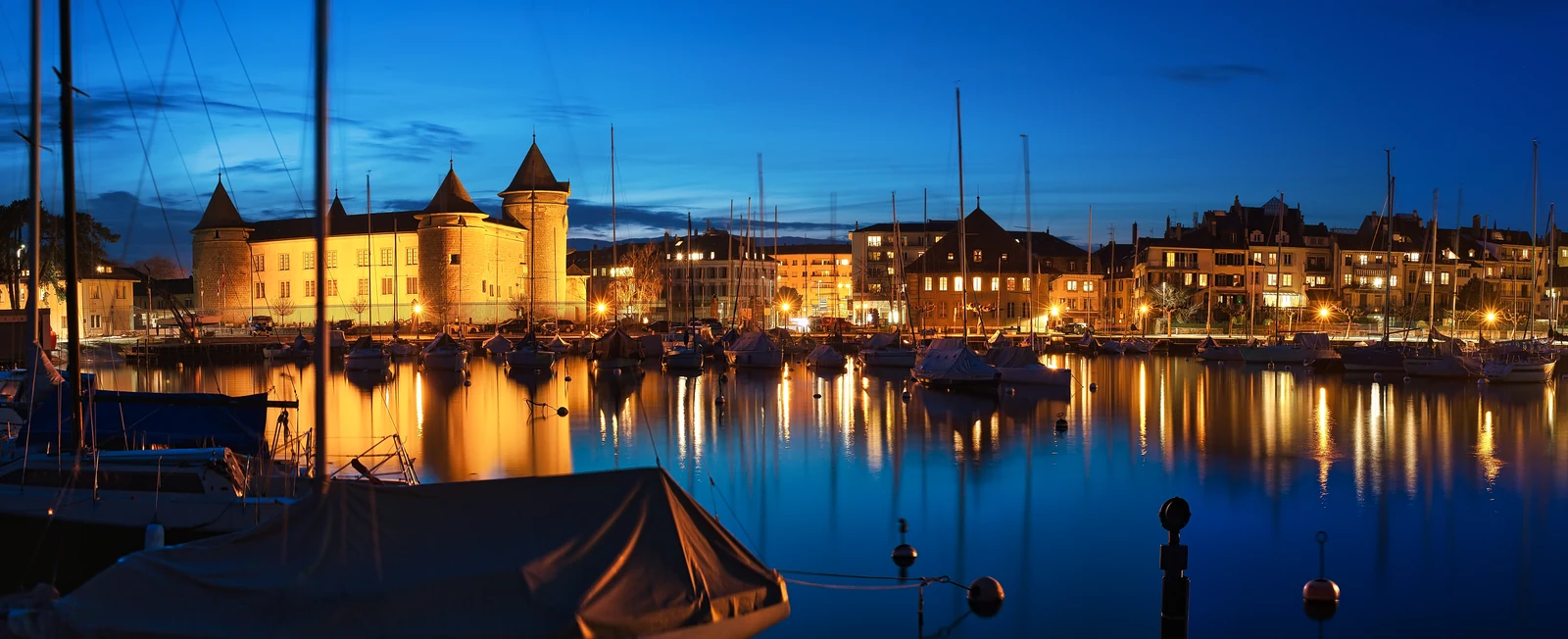Le château de Morges illuminé et les bateaux du vieux port à la tombée du jour.